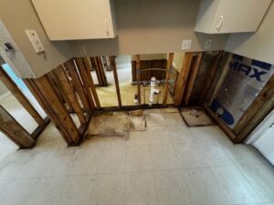 A room with lower wall sections removed, exposing wooden studs and plumbing pipes. The floor has white tiles, and upper wall cabinets remain intact. Some water damage and mold are visible on the studs and floor edges.