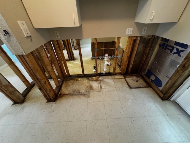A room with lower wall sections removed, exposing wooden studs and plumbing pipes. The floor has white tiles, and upper wall cabinets remain intact. Some water damage and mold are visible on the studs and floor edges.