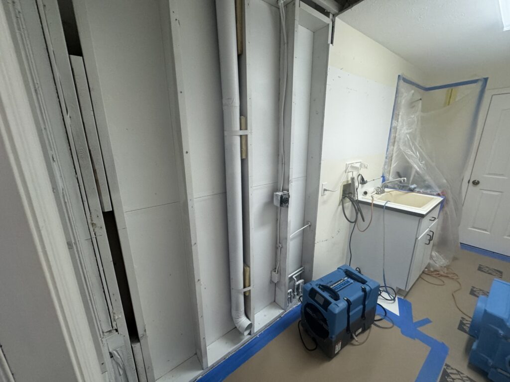 A partially renovated room with exposed pipes and wall studs, a blue air mover on the floor, taped protective coverings, a small sink, and construction materials near a closed white door.