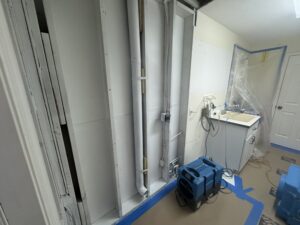 A partially renovated room with exposed pipes and wall studs, a blue air mover on the floor, taped protective coverings, a small sink, and construction materials near a closed white door.