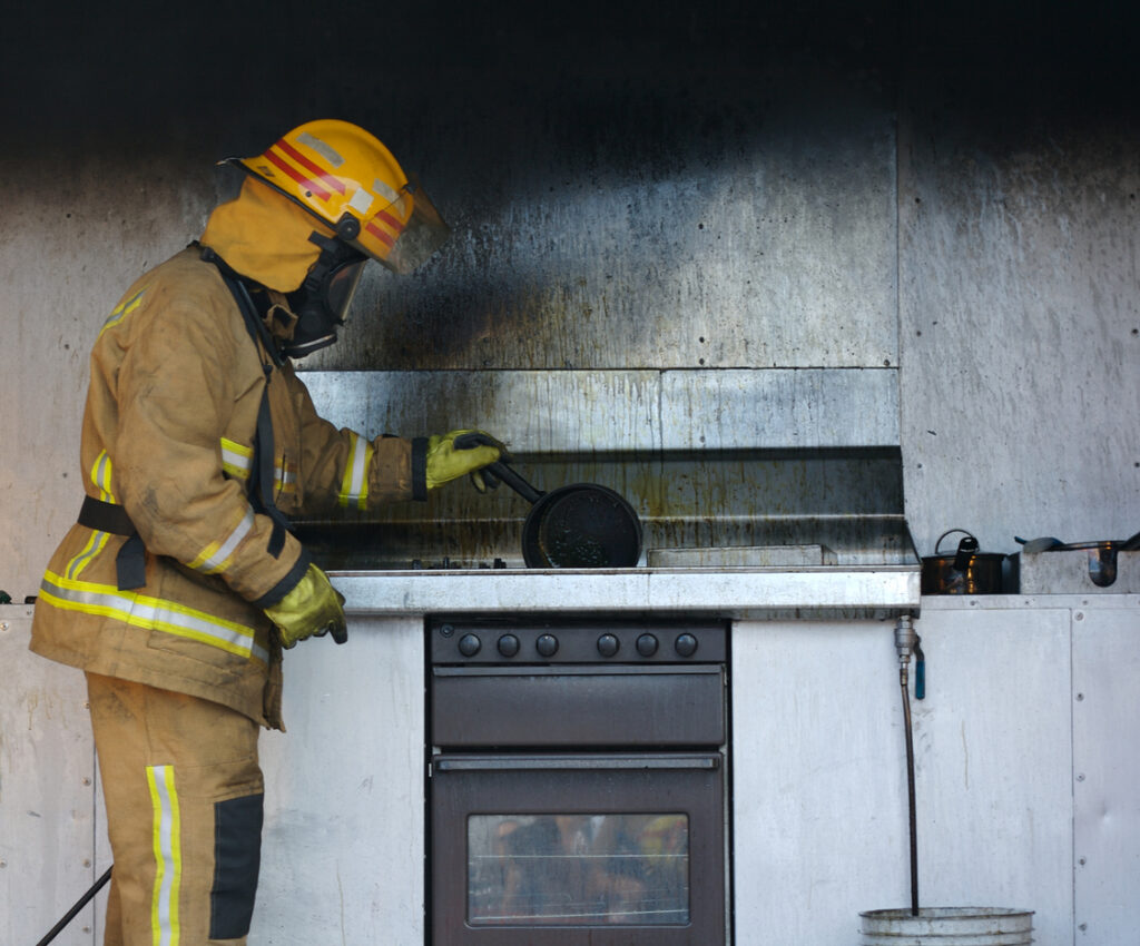 A firefighter in full gear inspects a burnt stovetop and wall in a kitchen, holding a pan, with soot and smoke damage visible on the metal backsplash.