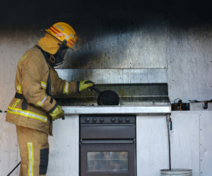 A firefighter in full gear inspects a burnt stovetop and wall in a kitchen, holding a pan, with soot and smoke damage visible on the metal backsplash.